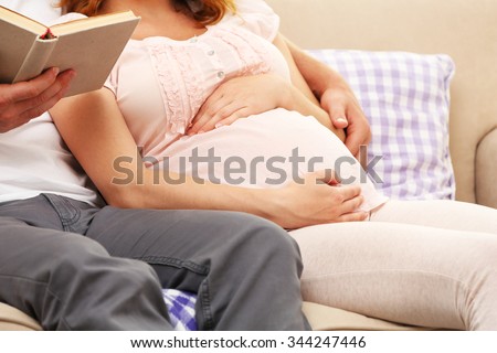 Handsome man reads book to his lovely pregnant woman on sofa in the room, close up