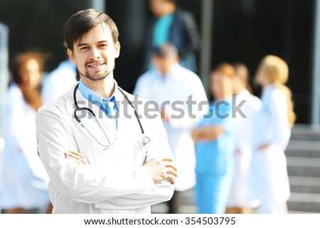 Smiling doctor with medical stuff behind standing against clinic entrance