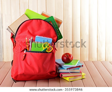 Full of stationary red backpack and pile of books with apple on top on wooden table