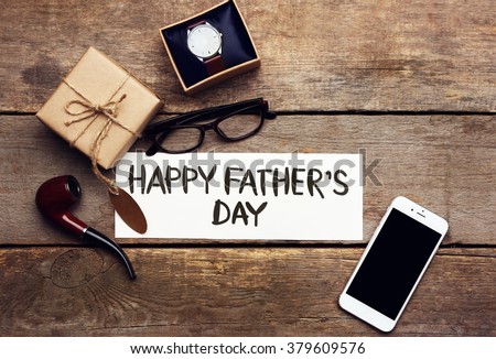 Happy Father\'s Day inscription with mobile phone and pipe on wooden background. Greetings and presents