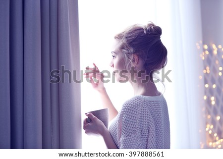 Blonde beautiful  girl looking in the window and holding a cup of coffee or tea in her hands