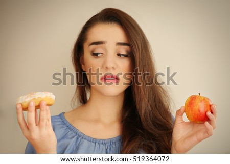 Beautiful young woman making choice between apple and donut on light background