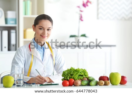 Young female nutritionist sitting at table with fresh vegetables and fruits in her office