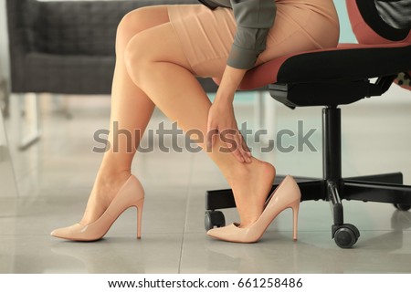 Young woman suffering from leg pain in office because of uncomfortable shoes