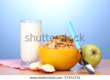 tasty cornflakes in yellow bowl, apples and glass of milk on wooden table on blue background