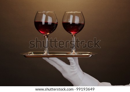 Hand in glove holding silver tray with wineglasses on brown background