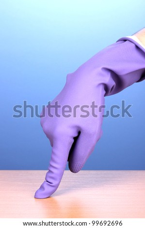Hand in color cleaning glove holding finger over surface of wooden table on blue background