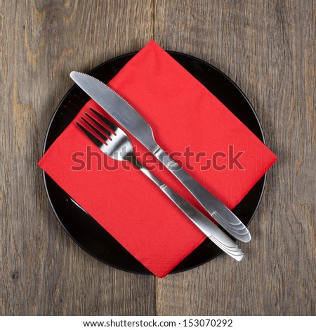Fork, knife and red napkin on black plate. Wooden background. Square format.