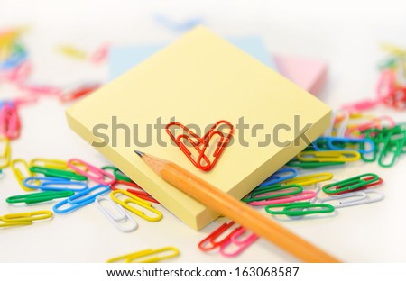Small note pad and a pencil on a background of colored paper clips on white.
