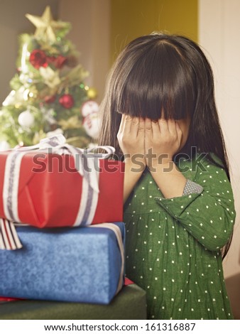 little asian girl covering her eyes in front of christmas gifts.