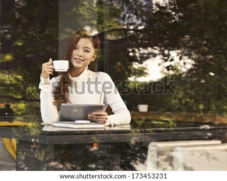young woman sitting next to windows drinking coffee with tablet computer in hand in cafe.