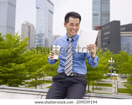 asian businessman looking at tablet holding coffee cup smiling.