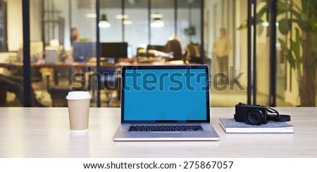 a laptop computer with blue screen, together with a camera and a cup of coffee on desk in a small stylish office.