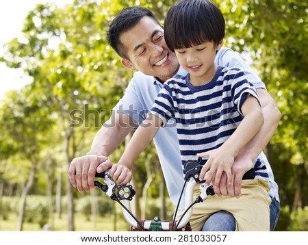 asian father and elementary-age son enjoying riding a bike outdoors in a park.