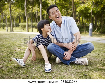 asian father and elementary-age son sitting on grass outdoors having an interesting conversation.