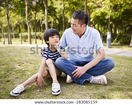 asian father and elementary-age son sitting on grass outdoors having a conversation.