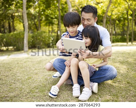 asian father and two children sitting on grass looking at tablet computer, outdoor in a park.