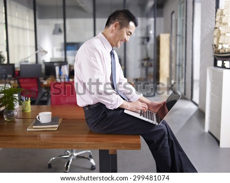 an asian businessman sitting on desk using laptop computer in office, happy and smiling, side view.