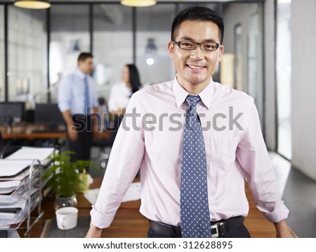 asian businessman standing in office happy and smiling with multinational colleagues talking in background.