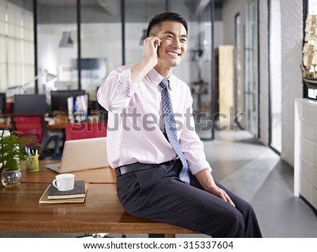 asian business person sitting on desk talking on mobile phone in office.
