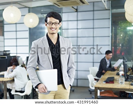 young asian entrepreneur standing in office with laptop computer under arm.