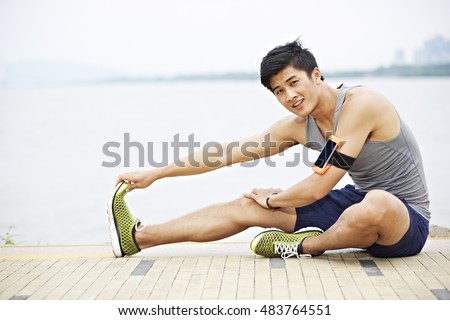 young handsome asian jogger wearing fitness tracker stretching a leg by lake looking at camera smiling