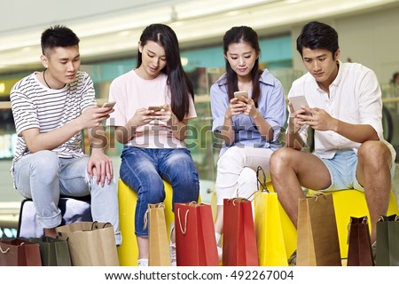 a group of young asian people men and women playing with cellphones while taking a break in shopping mall