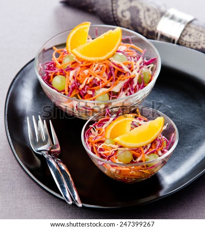 salad with red cabbage, carrots orange grapes in a glass bowl on a black plate with a napkin in the background