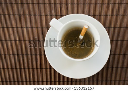 Cup of coffee with the cigarette inside, composition over a straw mat background