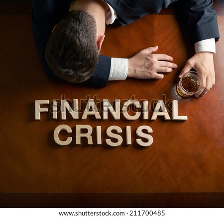 Phrase Financial Crisis made of wooden block letters and devastated caucasian man in a black suit sitting at the table with the glass of whiskey, top view composition with dramatic lighting