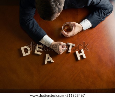 Word Death made of wooden block letters and devastated middle aged caucasian man in a black suit sitting at the table with the glass of whiskey, top view composition with dramatic lighting