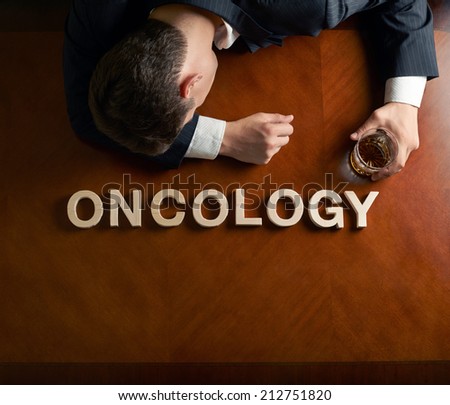 Word Oncology made of wooden block letters and devastated middle aged caucasian man in a black suit sitting at the table with the glass of whiskey, top view composition with dramatic lighting