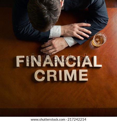 Phrase Financial Crime made of wooden block letters and devastated middle aged caucasian man in a black suit sitting at the table with the glass of whiskey, top view composition with dramatic lighting