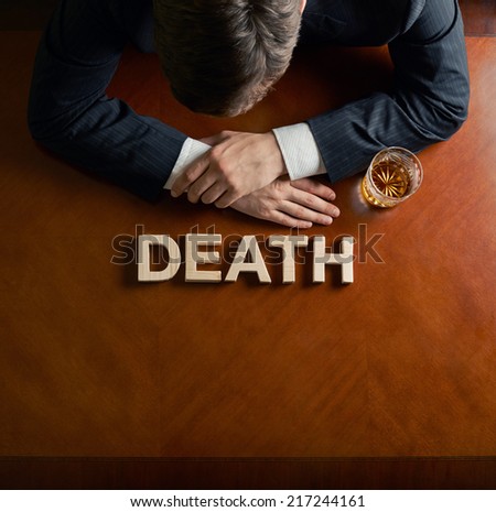 Word Death made of wooden block letters and devastated middle aged caucasian man in a black suit sitting at the table with the glass of whiskey, top view composition with dramatic lighting