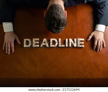 Word Deadline made of wooden block letters and devastated middle aged caucasian man in a black suit sitting at the table, top view composition with dramatic lighting