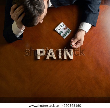 Word Pain made of wooden block letters and devastated middle aged caucasian man in a black suit sitting at the table, top view composition with dramatic lighting