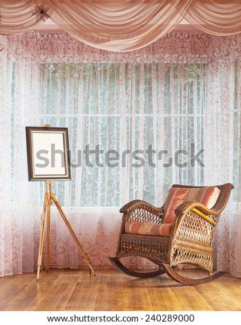 Copyspace empty picture frame on the wooden easel and next to wicker rocking chair, composition against the window\'s curtains background