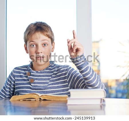 Studying 12 years old children boy sitting at the wooden desk over a book, looking to the camera with his pointing finger up, composition against the window