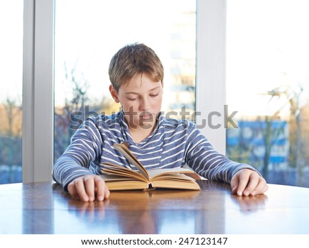 12 years old children boy sitting at the wooden desk while studying and reading a book, composition against the window