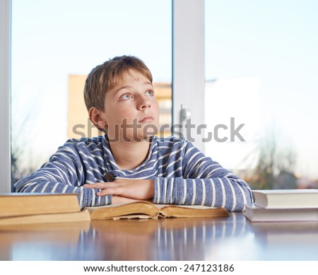 Dreamy 12 years old children boy sitting at the wooden desk over the book, composition against the window