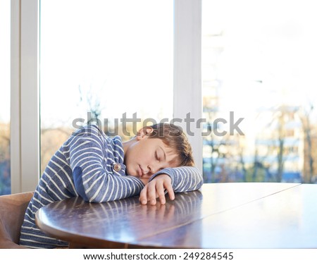 Sleepy 12 years old children boy sitting at the wooden desk, composition against the window