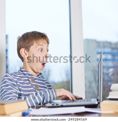12 years old children boy sitting at the wooden desk while playing with the notebook computer, composition against the window