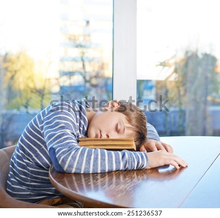Sleepy 12 years old children boy sitting at the wooden desk and sleeping over the book, composition against the window