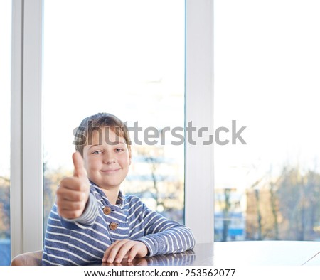 Happy 12 years old children boy sitting at the wooden desk showing a thumbs up gesture, composition against the window