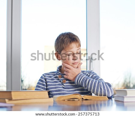 12 years old childen boy sitting at the wooden desk while studying and reading a book, composition against the window