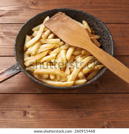 Cooking fried french potatoes composition of an old iron pan, wooden spatula and pile of fries over the wooden table\'s surface