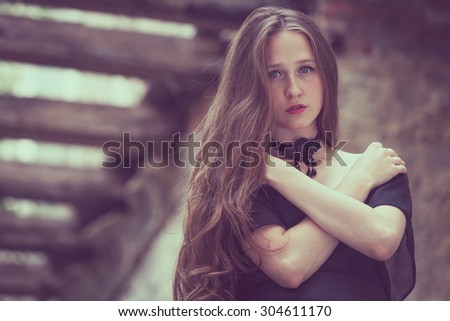 portrait of a beautiful young sad goth girl in a black dress standing in an abandoned old building outdoors at the day time