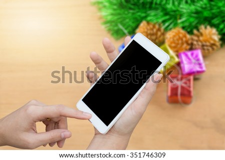 woman hand hold and touch smart phone,cell phone,mobile over blurred image of christmas decoration,gift sitting on a table with a tree in the background