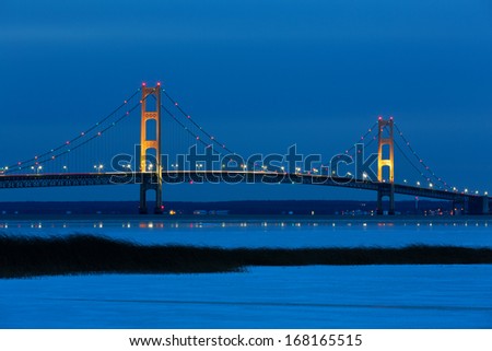 Blue Hour, just after sunset, creates an inviting backdrop for the Mackinac Bridge. Its lights twinkle and create reflections on winter ice. Snow and grass in the foreground add to this fine art photo