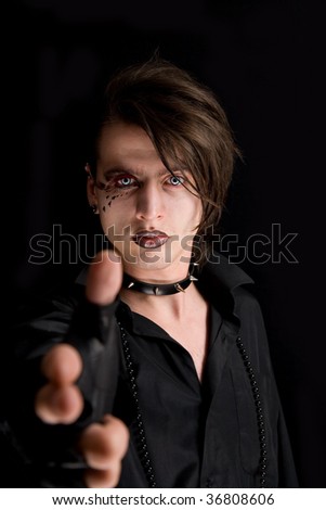 Gothic boy with artistic make-up pointing his hand as a gun, isolated on black background